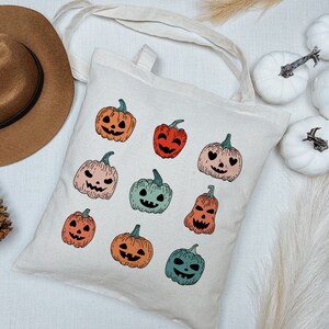 May include: A white canvas tote bag with a variety of colorful jack-o'-lantern designs. The bag has a brown leather handle and is perfect for carrying everyday essentials.