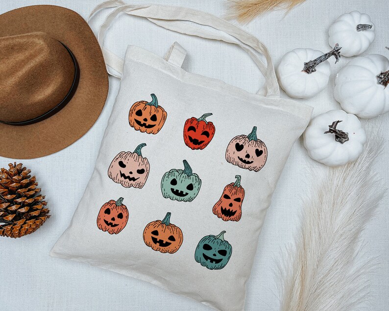 May include: A white canvas tote bag with a variety of colorful jack-o'-lantern designs. The bag has a brown leather handle and is perfect for carrying everyday essentials.