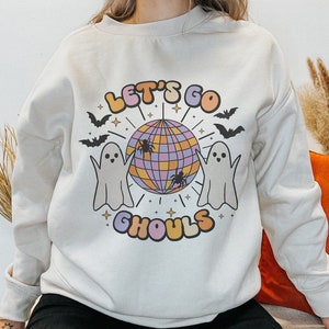 May include: A cream-colored sweatshirt with a graphic design featuring two ghosts dancing around a disco ball. The text "Let's Go Ghouls" is printed in a colorful font below the disco ball.