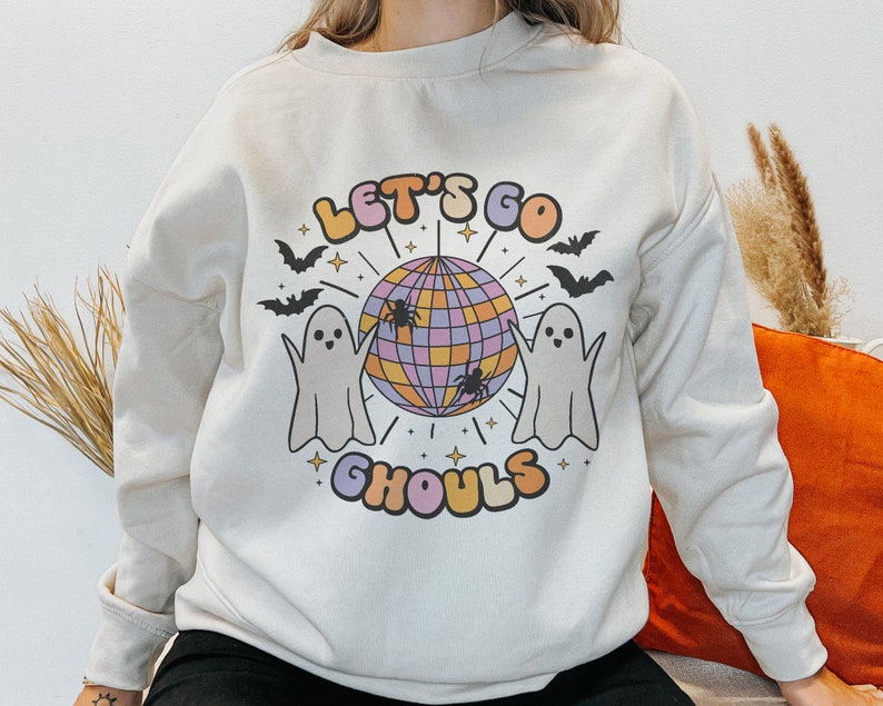 May include: A cream-colored sweatshirt with a graphic design featuring two ghosts dancing around a disco ball. The text "Let's Go Ghouls" is printed in a colorful font below the disco ball.