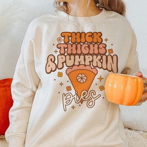 K&ouml;nnte beinhalten: Ein cremefarbener Pullover mit dem Text "Thick Thighs & Pumpkin Pies" in einer Retro-Schrift. Der Text ist von kleinen orangefarbenen Blumen und Sternen umgeben. Eine Person h&auml;lt eine Tasse in Form eines K&uuml;rbisses.