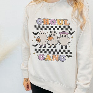 Puede incluir: Una sudadera blanca con un gr&aacute;fico de tres fantasmas en l&iacute;nea. Los fantasmas llevan sombreros y llevan calabazas. El texto "Ghoul Gang" est&aacute; impreso en letras de colores debajo de los fantasmas.