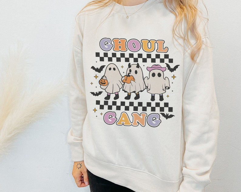 Puede incluir: Una sudadera blanca con un gr&aacute;fico de tres fantasmas en l&iacute;nea. Los fantasmas llevan sombreros y llevan calabazas. El texto "Ghoul Gang" est&aacute; impreso en letras de colores debajo de los fantasmas.