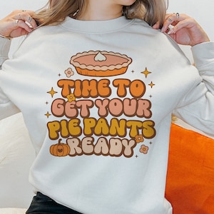 Pode incluir: Uma camisa de moletom branca com um design gr&aacute;fico retr&ocirc; que apresenta uma ab&oacute;bora, uma torta e o texto "Time to get your pie pants ready".