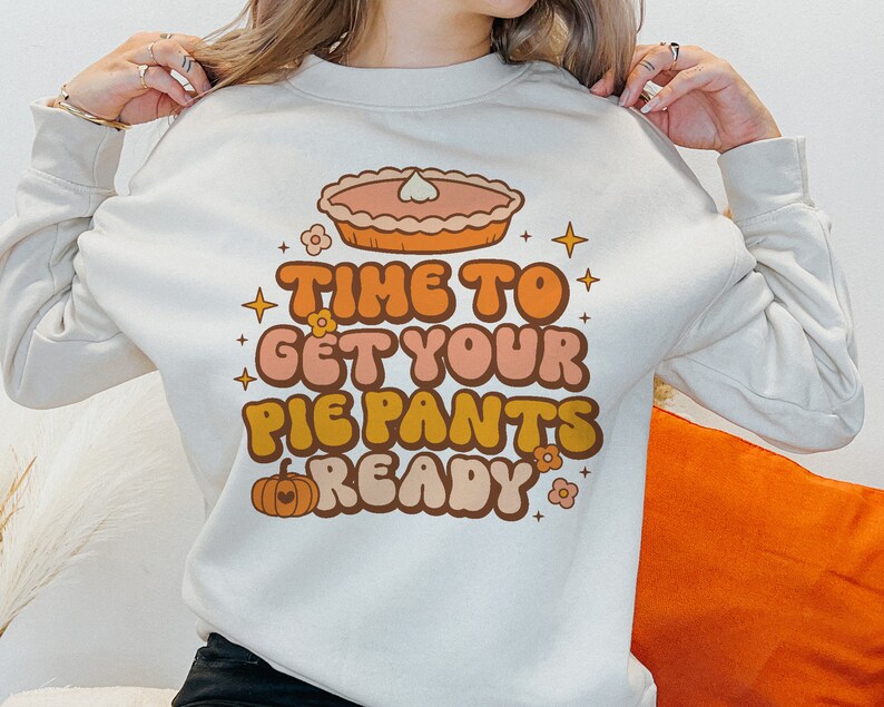 Pode incluir: Uma camisa de moletom branca com um design gr&aacute;fico retr&ocirc; que apresenta uma ab&oacute;bora, uma torta e o texto "Time to get your pie pants ready".