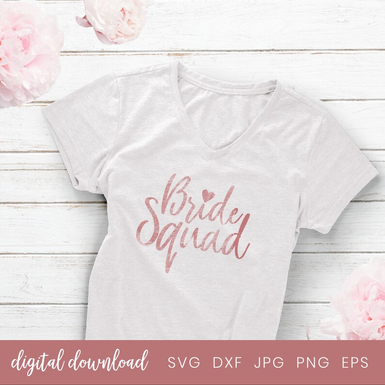 May include: A white t-shirt with a rose gold graphic that says "Bride Squad" with a small heart above the word "Bride".