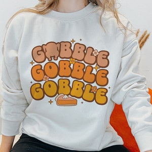 May include: A white sweatshirt with a graphic design of the words "Gobble Gobble Gobble" in a retro font with orange, yellow, and brown colors. The graphic also includes a small pie and stars.