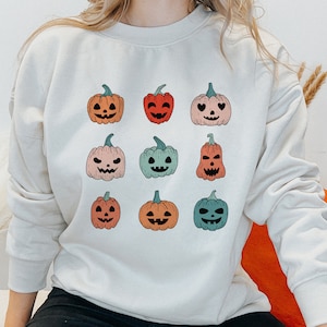 May include: A white sweatshirt with a design of nine colorful jack-o'-lantern pumpkins. The pumpkins have different facial expressions and are arranged in a grid pattern.