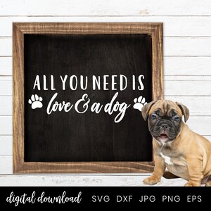 以下が含まれることがあります： 「ALL YOU NEED IS love & a dog」という白い文字が書かれた、枠付きの黒板の看板。文字の両側に肉球の跡があります。看板の前に、茶色と白の小さな子犬が座っています。