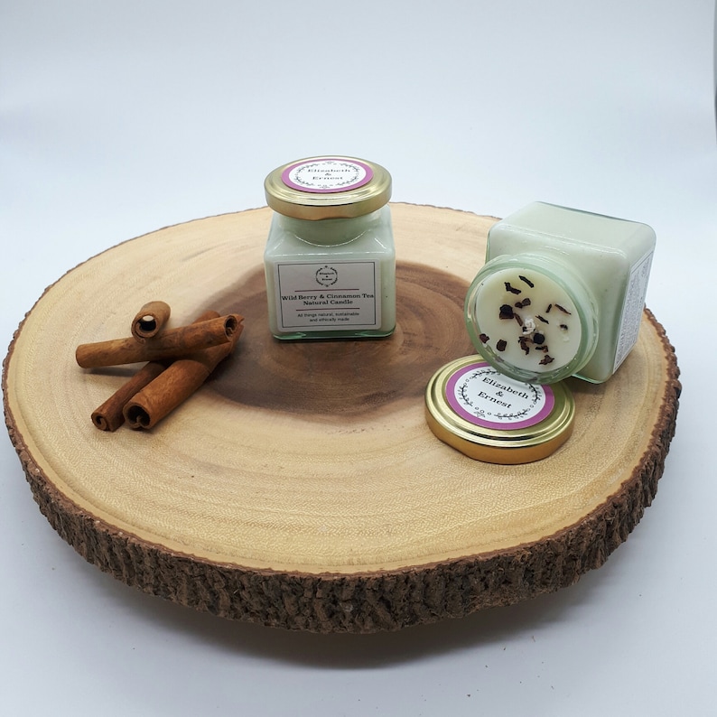 Może przedstawiać: Dwa małe szklane słoiki z pokrywkami zawierające naturalne świece sojowe. Świece są jasnozielone i mają etykietę z napisem "Wild Berry & Cinnamon Tea Natural Candle". Słoiki stoją na drewnianym plastrze z cynamonowymi patyczkami.