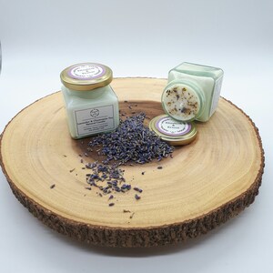 Może przedstawiać: Dwa małe szklane słoiki z pokrywkami zawierające naturalne świece sojowe. Świece są oznaczone etykietami "Lavender & Chamomile Tea Natural Candle" i "Elizabeth & Ernest". Słoiki stoją na drewnianym plastrze z rozrzuconymi wok&oacute;ł suszonymi kwiatami lawendy.