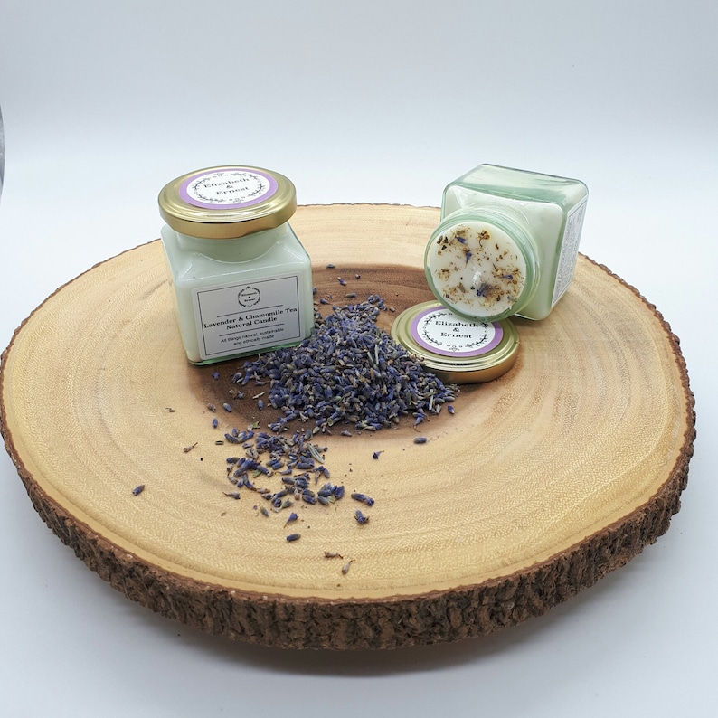 Może przedstawiać: Dwa małe szklane słoiki z pokrywkami zawierające naturalne świece sojowe. Świece są oznaczone etykietami "Lavender & Chamomile Tea Natural Candle" i "Elizabeth & Ernest". Słoiki stoją na drewnianym plastrze z rozrzuconymi wok&oacute;ł suszonymi kwiatami lawendy.