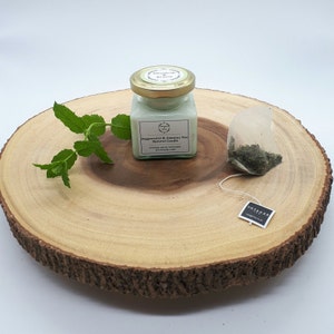 Może przedstawiać: Mała szklana świeca w słoiku z zieloną pokrywką i białą etykietą z napisem "Peppermint & Jasmine Tea Natural Candle". Świeca stoi na okrągłym drewnianym plastrze z zielonym listkiem mięty i saszetką herbaty.
