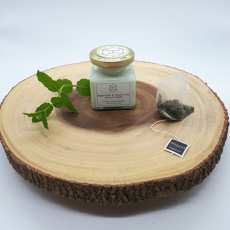Może przedstawiać: Mała szklana świeca w słoiku z zieloną pokrywką i białą etykietą z napisem "Peppermint & Jasmine Tea Natural Candle". Świeca stoi na okrągłym drewnianym plastrze z zielonym listkiem mięty i saszetką herbaty.