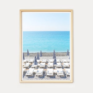 Puede incluir: Una escena de playa con tumbonas blancas y sombrillas a rayas azules y blancas en una playa de guijarros. El mar es de un azul turquesa brillante y el cielo es de un azul claro.