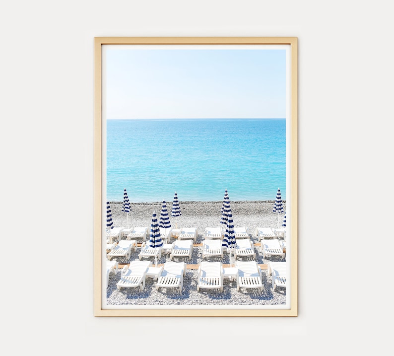 Puede incluir: Una escena de playa con tumbonas blancas y sombrillas a rayas azules y blancas en una playa de guijarros. El mar es de un azul turquesa brillante y el cielo es de un azul claro.