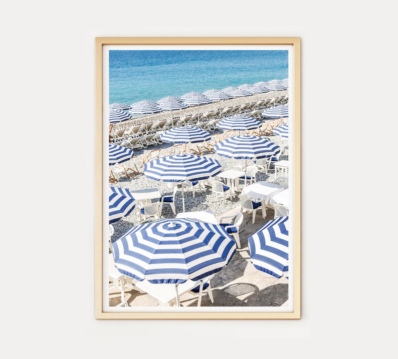 Puede incluir: Una escena de playa con sombrillas a rayas azules y blancas y sillas blancas en una playa de guijarros. Las sombrillas est&aacute;n dispuestas en filas y hay un mar azul claro en el fondo.