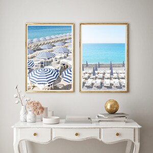 Puede incluir: Dos fotograf&iacute;as enmarcadas de una escena de playa con sombrillas a rayas azules y blancas y tumbonas blancas. La playa est&aacute; cubierta de peque&ntilde;as piedras. Las fotos est&aacute;n colgadas en una pared blanca sobre una c&oacute;moda blanca.