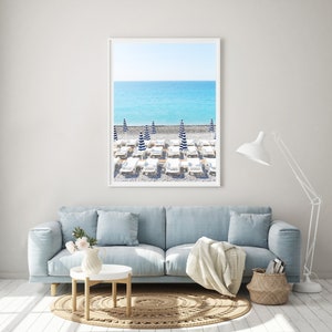 Puede incluir: Una fotografía enmarcada de una escena de playa con tumbonas blancas y sombrillas a rayas azules y blancas. La playa está cubierta de guijarros blancos y el océano es de un azul brillante.