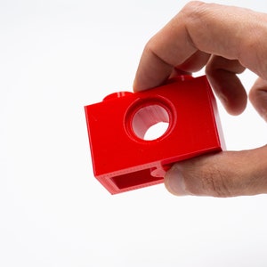 May include: A red plastic building block with a hole in the center. The block has a smooth surface and is designed for connecting with other blocks.