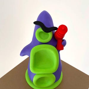 May include: A whimsical, handmade clay sculpture of a purple creature with green accents. It features a black mustache, green eye-like circles, and a red object held in its hand. The sculpture is set on a brown surface.