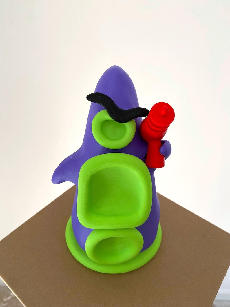 May include: A whimsical, handmade clay sculpture of a purple creature with green accents. It features a black mustache, green eye-like circles, and a red object held in its hand. The sculpture is set on a brown surface.