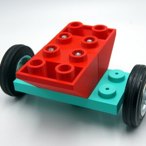 Puede incluir: Un ladrillo Lego rojo con seis tacos en la parte superior y cuatro tacos en los lados est&aacute; unido a una base Lego turquesa con dos ruedas negras.