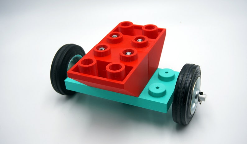 Puede incluir: Un ladrillo Lego rojo con seis tacos en la parte superior y cuatro tacos en los lados est&aacute; unido a una base Lego turquesa con dos ruedas negras.