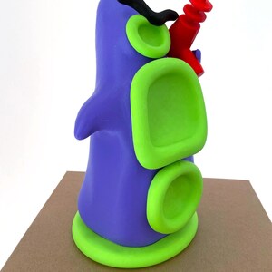 May include: A whimsical, abstract sculpture in shades of purple and green, with a red element. The sculpture has a rounded form with circular and rectangular green accents. A black, curved shape and a red object are attached to the top.