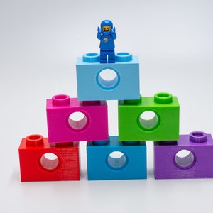 May include: A blue astronaut Lego figure stands on top of a stack of six colorful Lego blocks. The blocks are arranged in a pyramid shape, with the blue block at the top and the other blocks in a rainbow pattern.