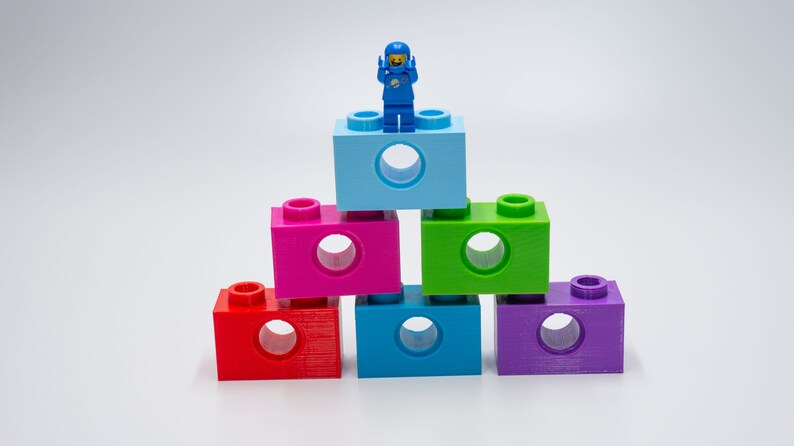 May include: A blue astronaut Lego figure stands on top of a stack of six colorful Lego blocks. The blocks are arranged in a pyramid shape, with the blue block at the top and the other blocks in a rainbow pattern.