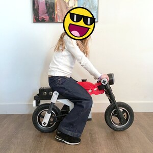 May include: A child sits on a red and black toy motorcycle with white accents. The motorcycle has two wheels and a handlebar.