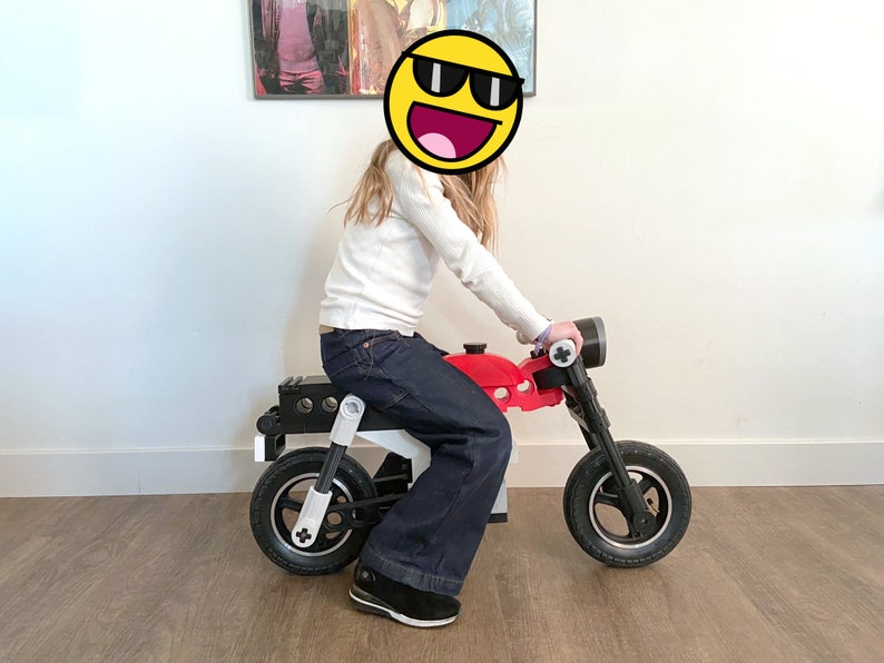 May include: A child sits on a red and black toy motorcycle with white accents. The motorcycle has two wheels and a handlebar.