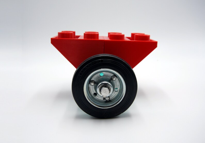 Puede incluir: Un carro de juguete rojo con cuatro ruedas. El carro est&aacute; hecho de bloques de construcci&oacute;n de pl&aacute;stico y tiene una rueda de goma negra con un cubo plateado.