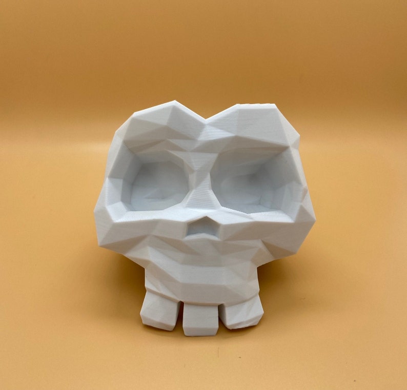 Op de afbeelding: Witte, geometrisch ontworpen schedel plantenpot. De schedel heeft een uitgeholde oogkas en een kleine, open mond.