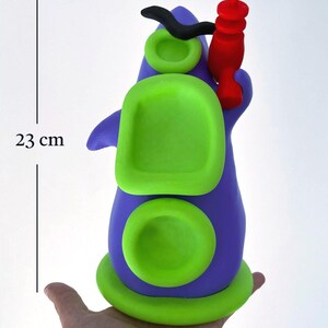 May include: A whimsical, handmade sculpture in shades of purple and green, with a red accent. The sculpture is approximately 9 inches tall. The design features rounded shapes and a unique, artistic aesthetic. The sculpture is held in a hand.