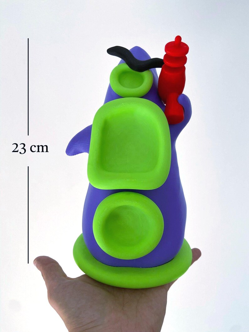 May include: A whimsical, handmade sculpture in shades of purple and green, with a red accent. The sculpture is approximately 9 inches tall. The design features rounded shapes and a unique, artistic aesthetic. The sculpture is held in a hand.