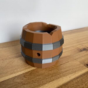 May include: A brown and gray 3D printed barrel with a hole in the side. The barrel is sitting on a wooden surface.