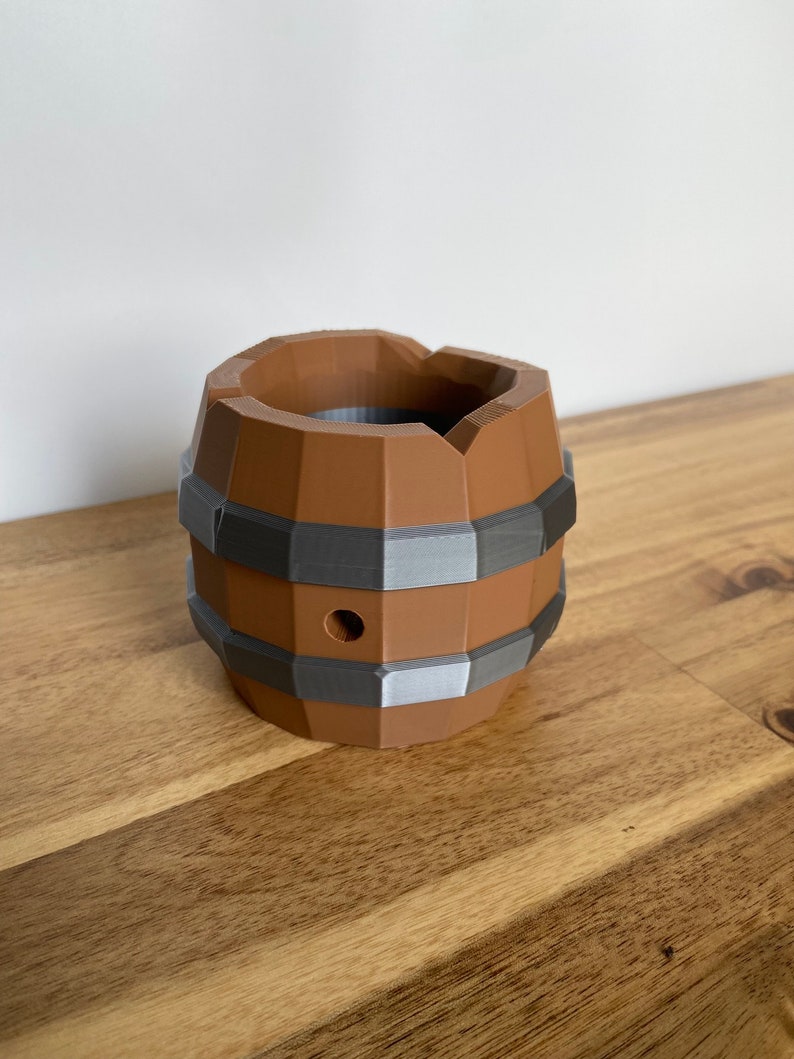May include: A brown and gray 3D printed barrel with a hole in the side. The barrel is sitting on a wooden surface.