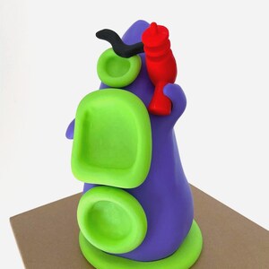 May include: A whimsical, abstract sculpture in shades of purple, green, black, and red. The form features rounded shapes and a stylized design, possibly representing a creature or character. The sculpture is placed on a brown surface.