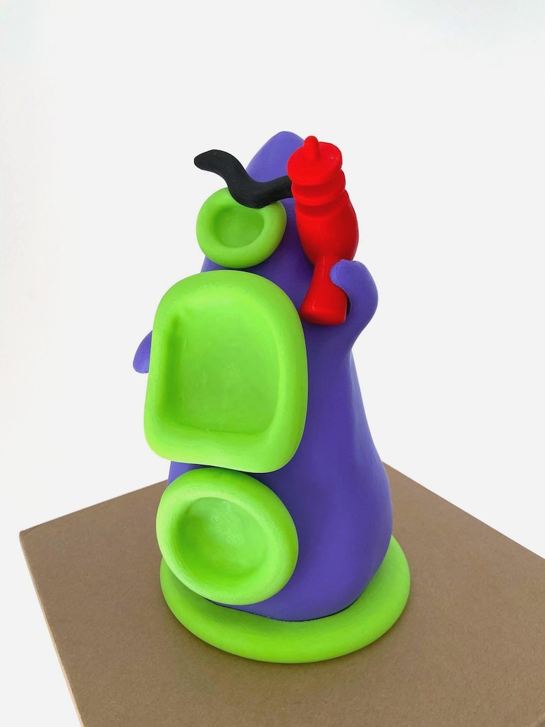 May include: A whimsical, abstract sculpture in shades of purple, green, black, and red. The form features rounded shapes and a stylized design, possibly representing a creature or character. The sculpture is placed on a brown surface.