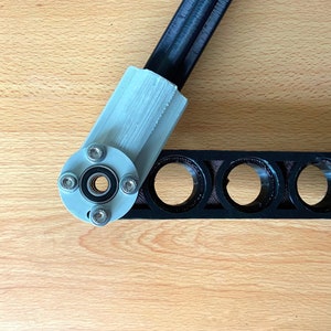 May include: A close-up of a black and silver 3D printed robotic arm joint. The joint has a bearing and is attached to a black plastic bar with multiple holes.