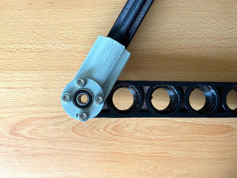 May include: A close-up of a black and silver 3D printed robotic arm joint. The joint has a bearing and is attached to a black plastic bar with multiple holes.