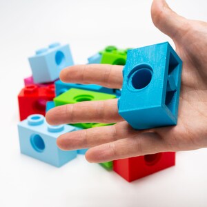 May include: A hand holding a blue plastic building block with a hole in the center. The block is surrounded by other colorful plastic building blocks.