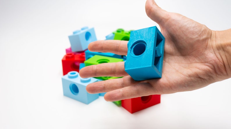 May include: A hand holding a blue plastic building block with a hole in the center. The block is surrounded by other colorful plastic building blocks.