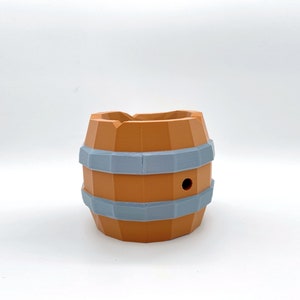 May include: A small, orange-brown ceramic planter shaped like a barrel with two gray bands around the middle. The planter has a small hole in the top.