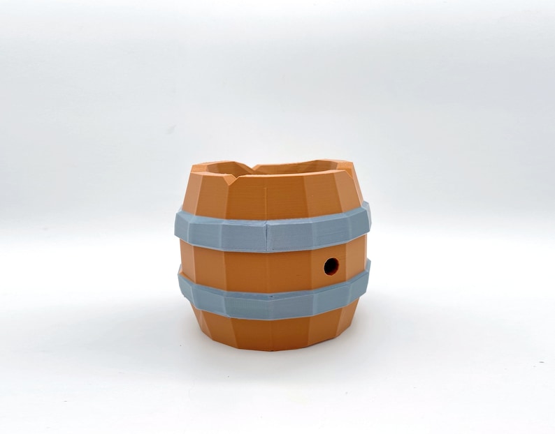 May include: A small, orange-brown ceramic planter shaped like a barrel with two gray bands around the middle. The planter has a small hole in the top.