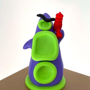May include: A whimsical, colorful sculpture of a purple creature with green accents and a red object. The sculpture has a rounded base, two circular green details, and a rectangular green shape. A black curved shape is on top.