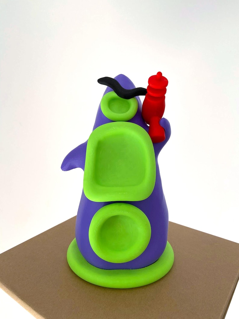 May include: A whimsical, colorful sculpture of a purple creature with green accents and a red object. The sculpture has a rounded base, two circular green details, and a rectangular green shape. A black curved shape is on top.