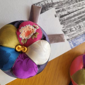 May include: A colorful, round, patchwork cushion with a floral embroidery. The cushion is made of various fabrics in shades of blue, yellow, pink, and purple. A gold-colored clasp secures the cushion. A second cushion is visible in the image.